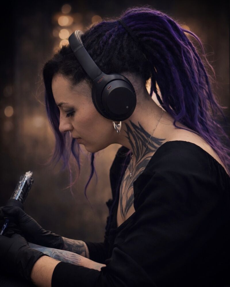 Kiara tatuando, de perfil, con los auriculares puestos y la máquina en la mano.